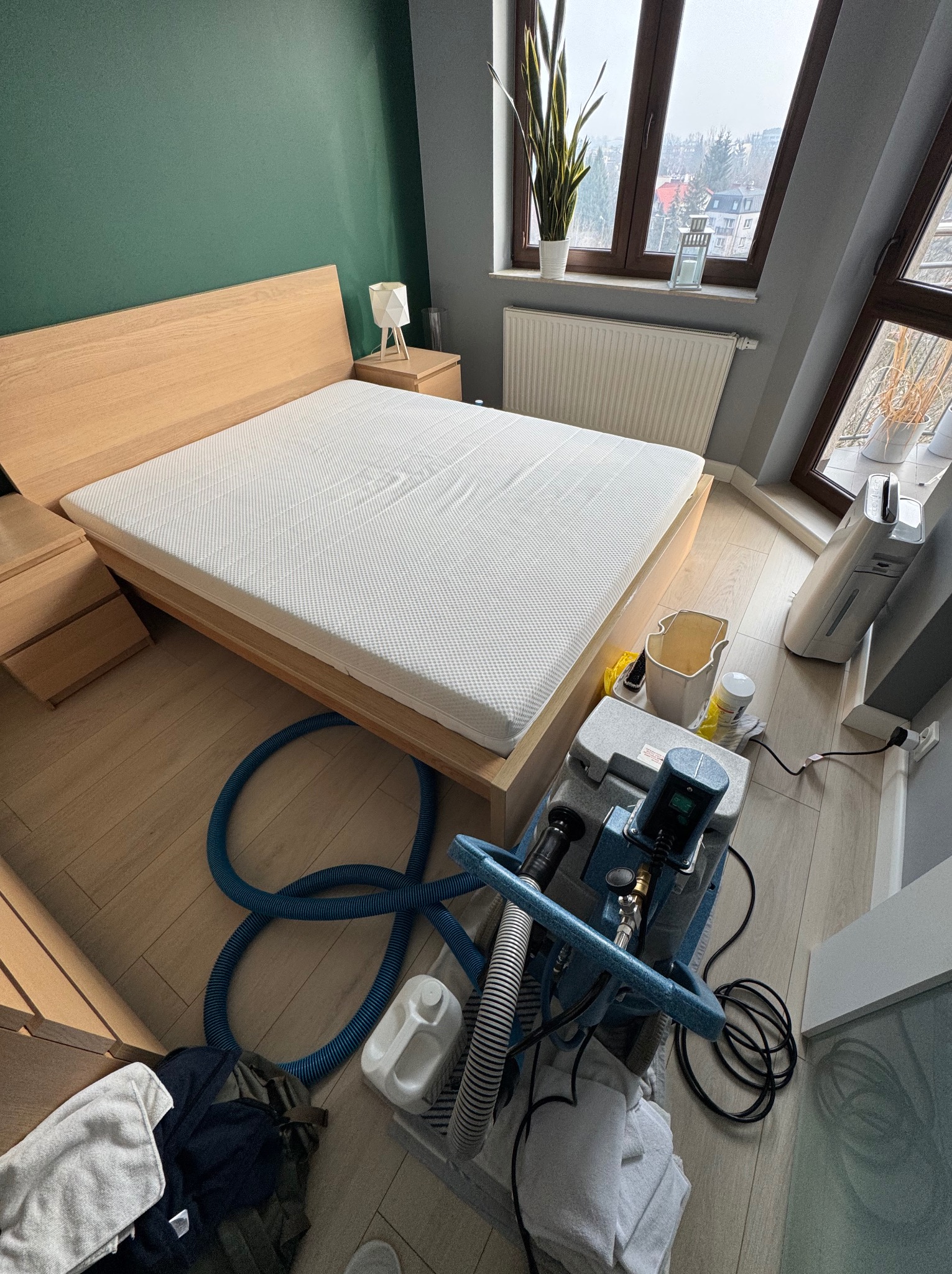 Jasne, drewniane łóżko z materacem w pokoju. Obok stoi sprzęt do czyszczenia tapicerki, sugerujący usługę prania materaca.