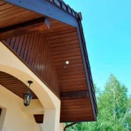 Drewniana podbitka dachowa z oświetleniem punktowym, widok z dołu na jasnym tle nieba i drzew, z elementami elewacji budynku.