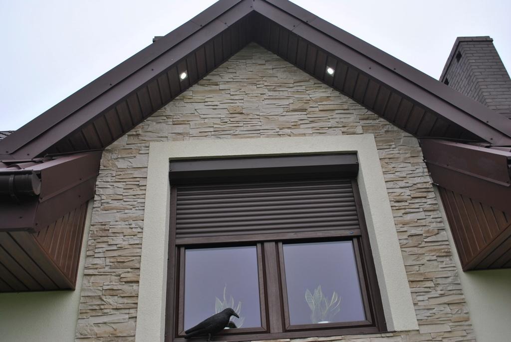 Dom z elewacją z kamienia, brązowymi rynnami i zamkniętą roletą okienną, na parapecie czarny ptak.