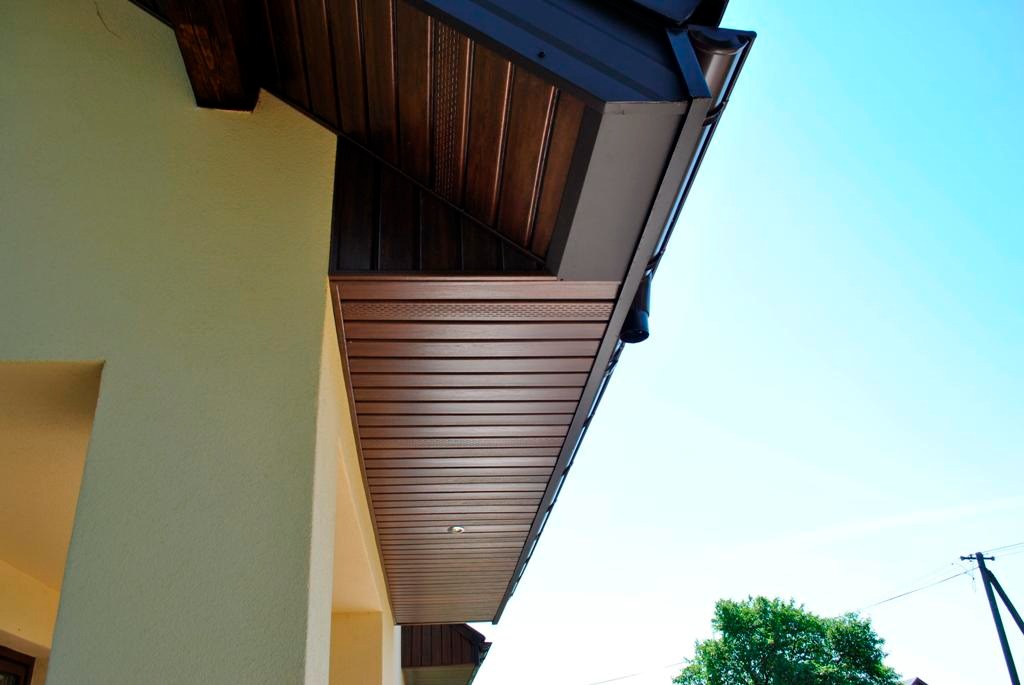 Nowoczesne podbitki dachowe z ciemnego drewna, widok z dołu, jasne niebo w tle, fragment rynny.