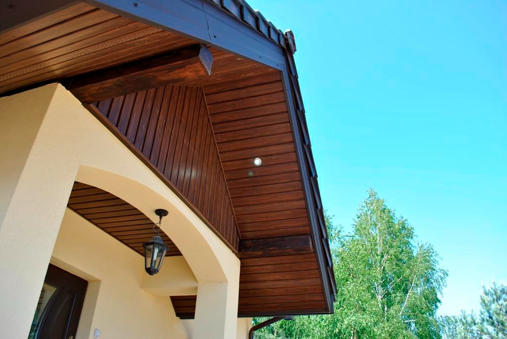 Drewniana podbitka dachowa z oświetleniem punktowym, widok z dołu na jasnym tle nieba i drzew, z elementami elewacji budynku.