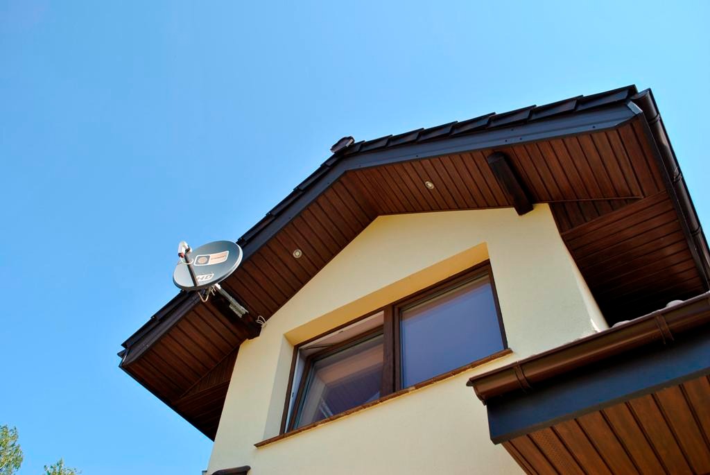 Fragment elewacji domu z brązowymi rynnami, drewnianą podbitką i anteną satelitarną na tle błękitnego nieba.