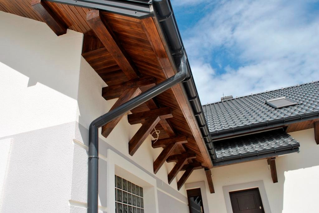 Czarna, metalowa rynna spustowa zamontowana na jasnej elewacji budynku z drewnianą podbitką i grafitowym dachem z dachówki ceramicznej.