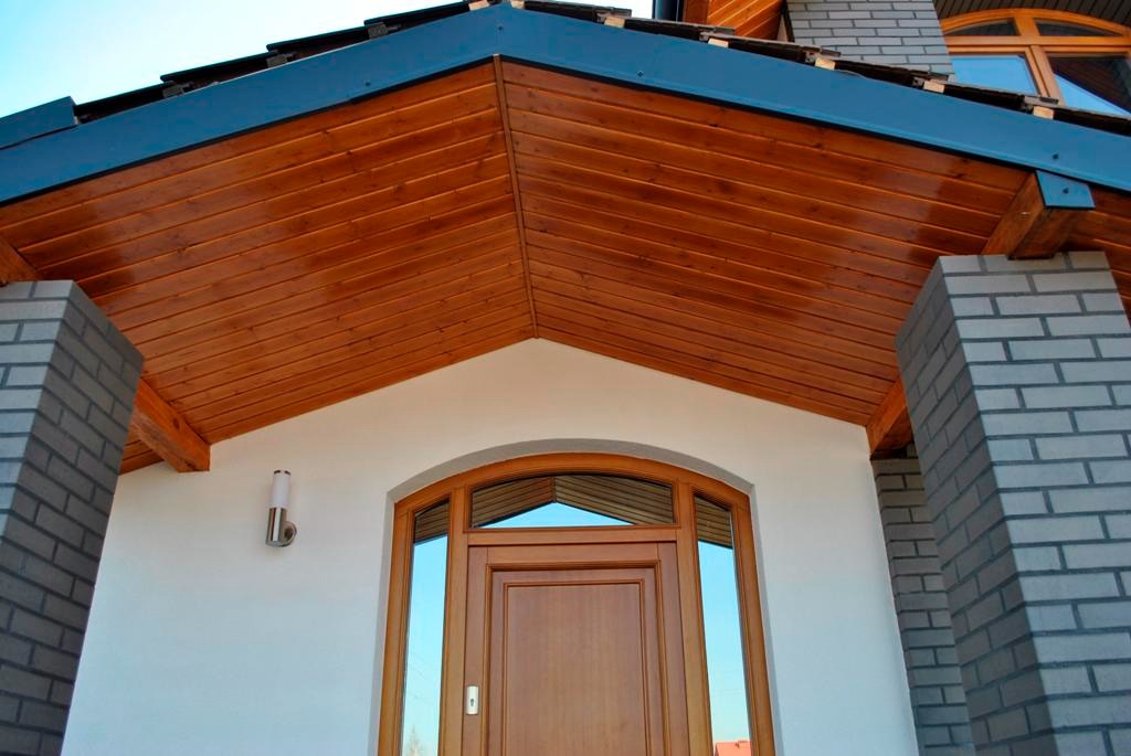 Drewniana podbitka dachowa z metalowym okapem nad wejściem z łukowymi oknami i drewnianymi drzwiami, oświetlenie zewnętrzne, ceglane filary.
