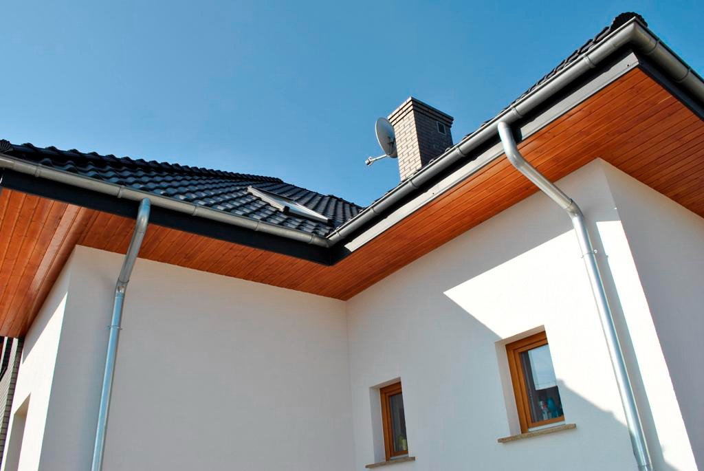 Nowoczesny dom jednorodzinny z ciemną dachówką i nowymi, metalowymi rynnami w słoneczny dzień. Widoczny fragment elewacji z dwoma oknami i drewnianą podbitką.