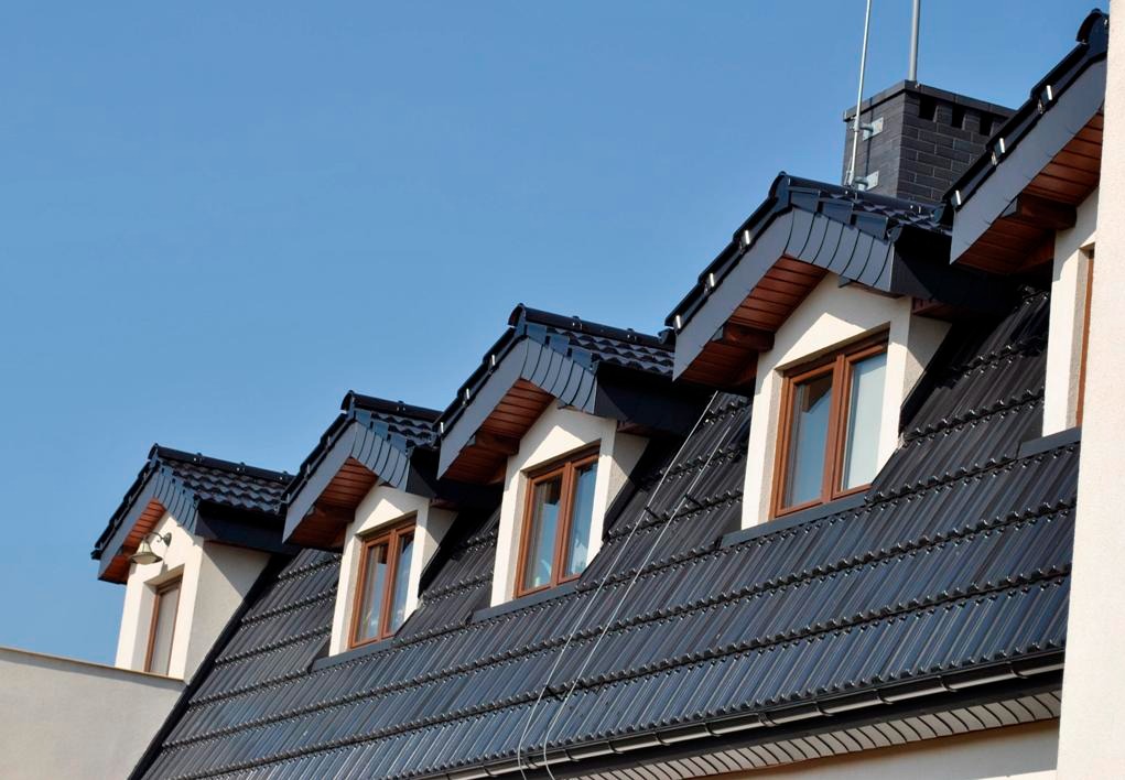 Dachówka karpiówka w kolorze antracytowym na dachu z oknami mansardowymi, widok z dołu na tle błękitnego nieba, z kominem w tle.