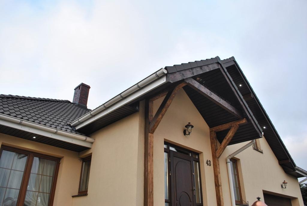 Dom jednorodzinny z ciemną dachówką, jasną elewacją i drewnianą konstrukcją wejściową z numerem 43, widoczne rynny w kolorze białym.