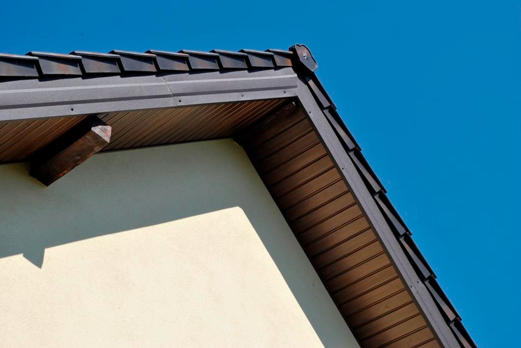 Szczegółowe ujęcie fragmentu dachu z brązową podbitką i ciemnoszarą obróbką blacharską, widoczne elementy mocowania, kremowa ściana budynku i błękitne niebo w tle.