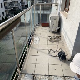 Pro bud compani - Balkon w bloku w trakcie remontu: usunięte płytki podłogowe, widoczne pozostałości kleju, zwinięty przedłużacz i klimatyzator na ścianie.