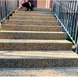 Pro bud compani - Schody zewnętrzne wykończone drobnym kamieniem w jasnych odcieniach, z czarną metalową barierką, widok z dołu, osoba w górnej części schodów wykonuje prace wykończeniowe.