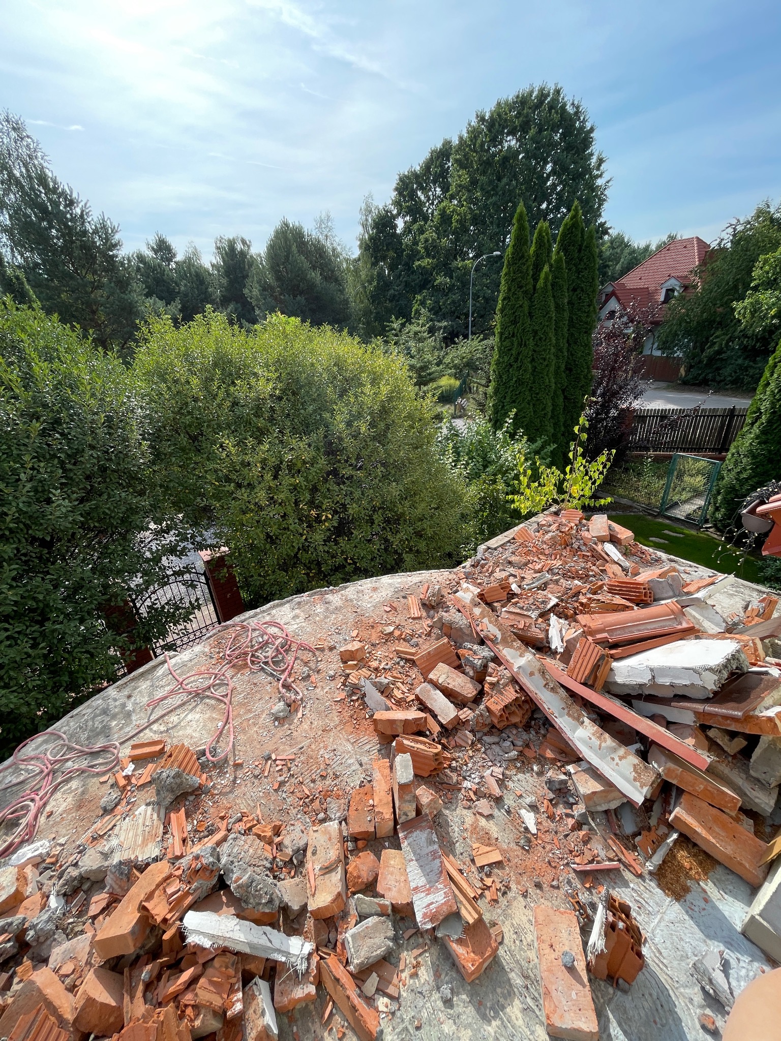 Widok z góry na gruz ceglany i fragmenty konstrukcji na betonowym podłożu, prawdopodobnie pozostałości po demontażu lub remoncie dachu z widocznym otoczeniem zieleni i zabudowań.