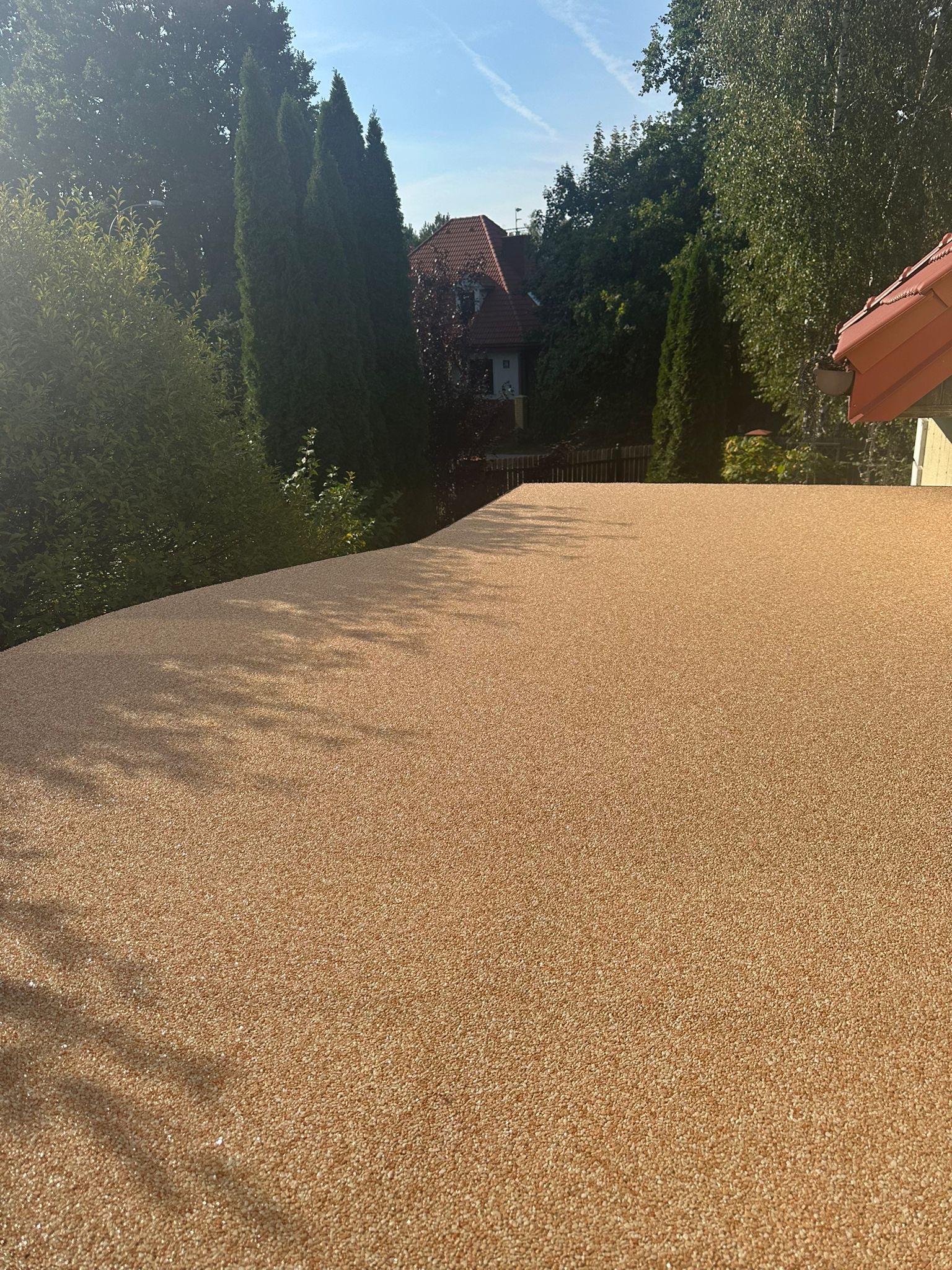 Beżowa, żywiczna powierzchnia balkonu z widocznymi cieniami drzew i fragmentem dachu sąsiedniego budynku w tle.