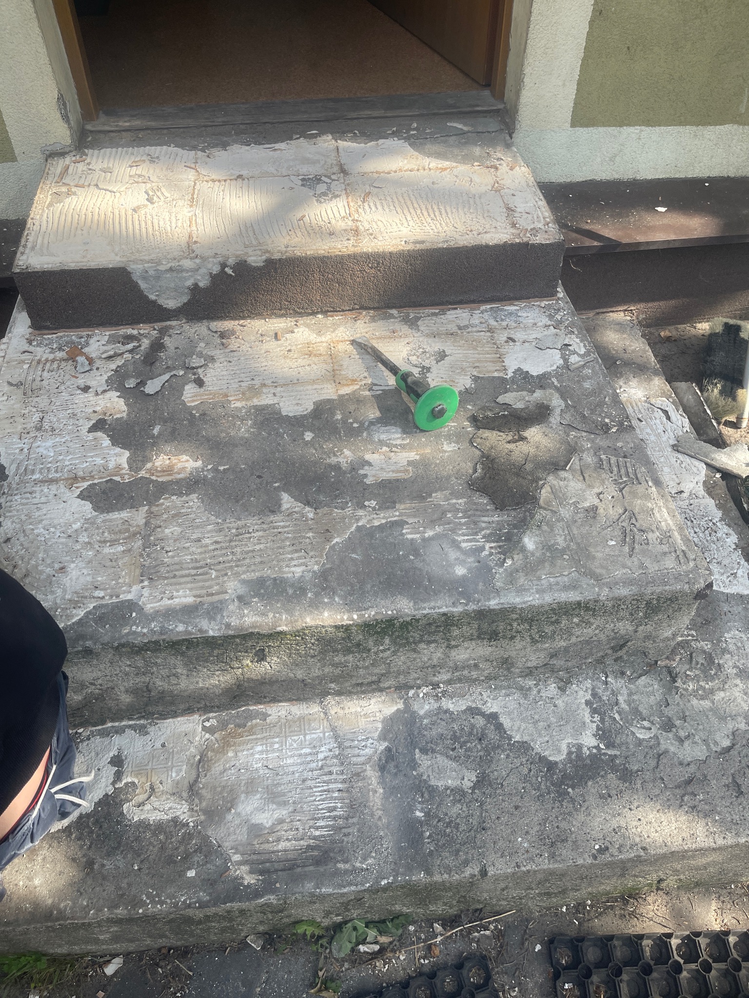 Schody wejściowe z usuniętymi płytkami, widoczne ślady po kleju i narzędzie do cięcia płytek z zieloną tarczą.