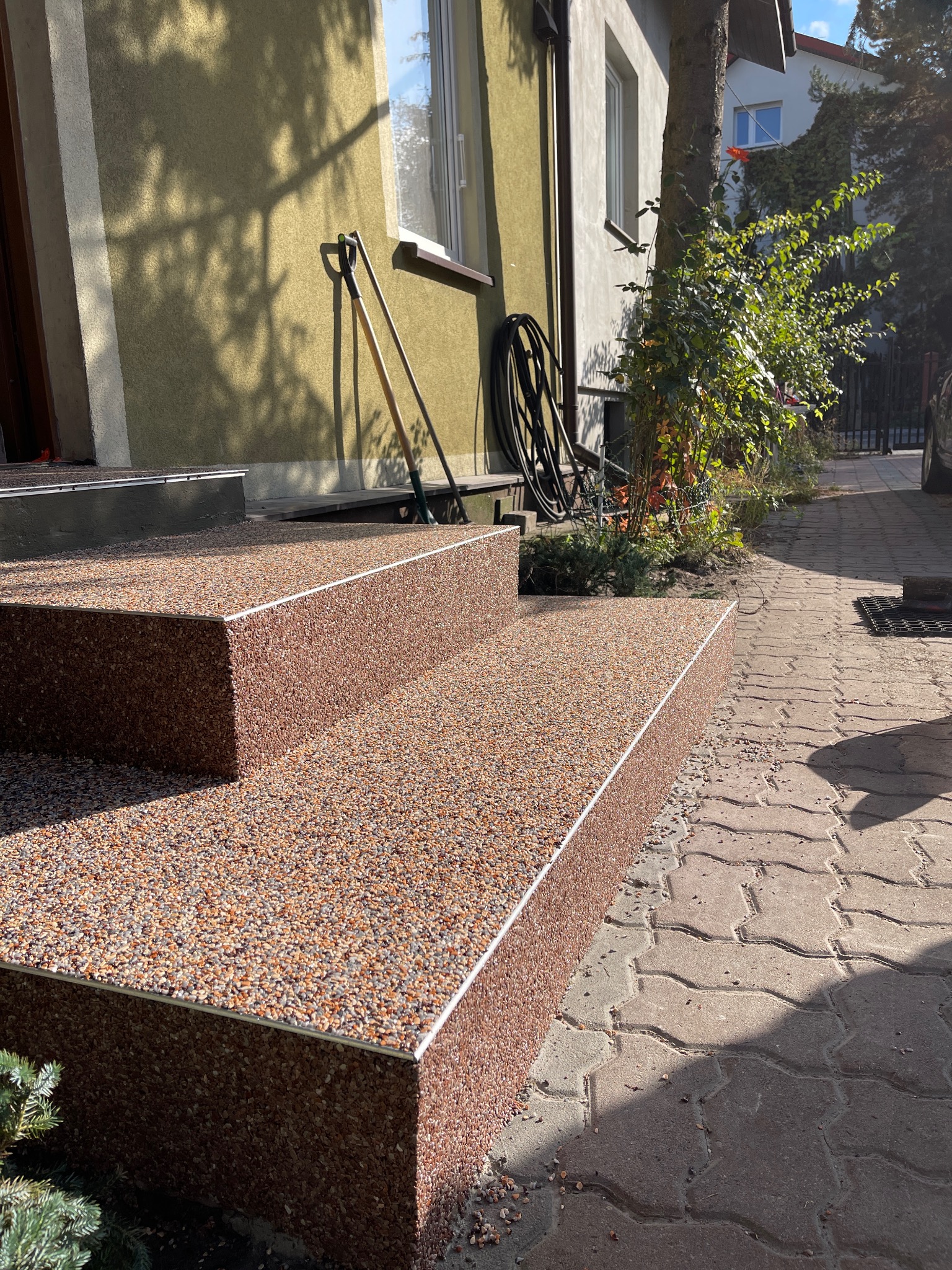 Schody wejściowe do domu wykończone żywicą epoksydową z kamyczkami, z metalowym obramowaniem krawędzi, na tle elewacji i chodnika z kostki brukowej.