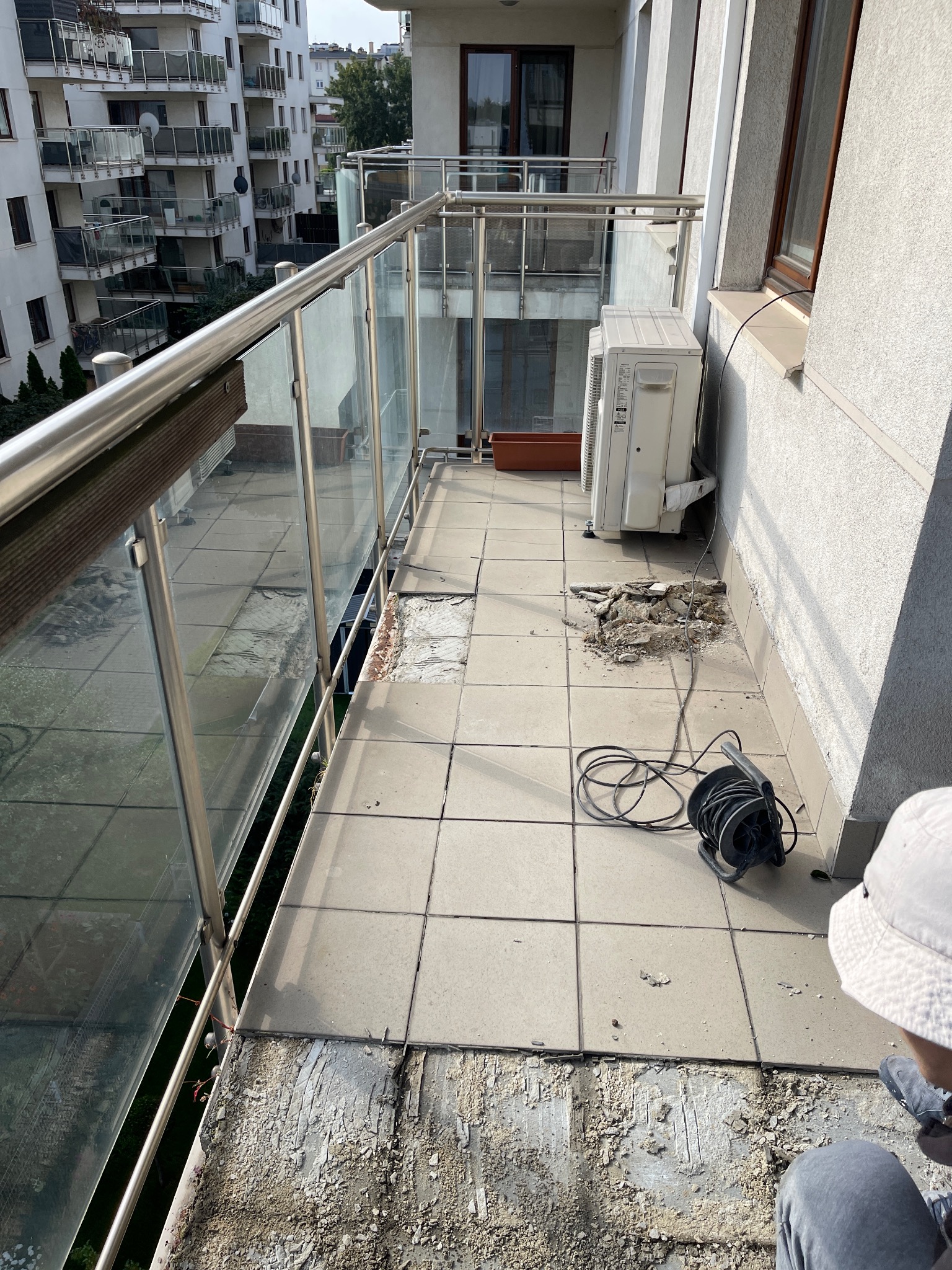 Balkon w bloku w trakcie remontu: usunięte płytki podłogowe, widoczne pozostałości kleju, zwinięty przedłużacz i klimatyzator na ścianie.