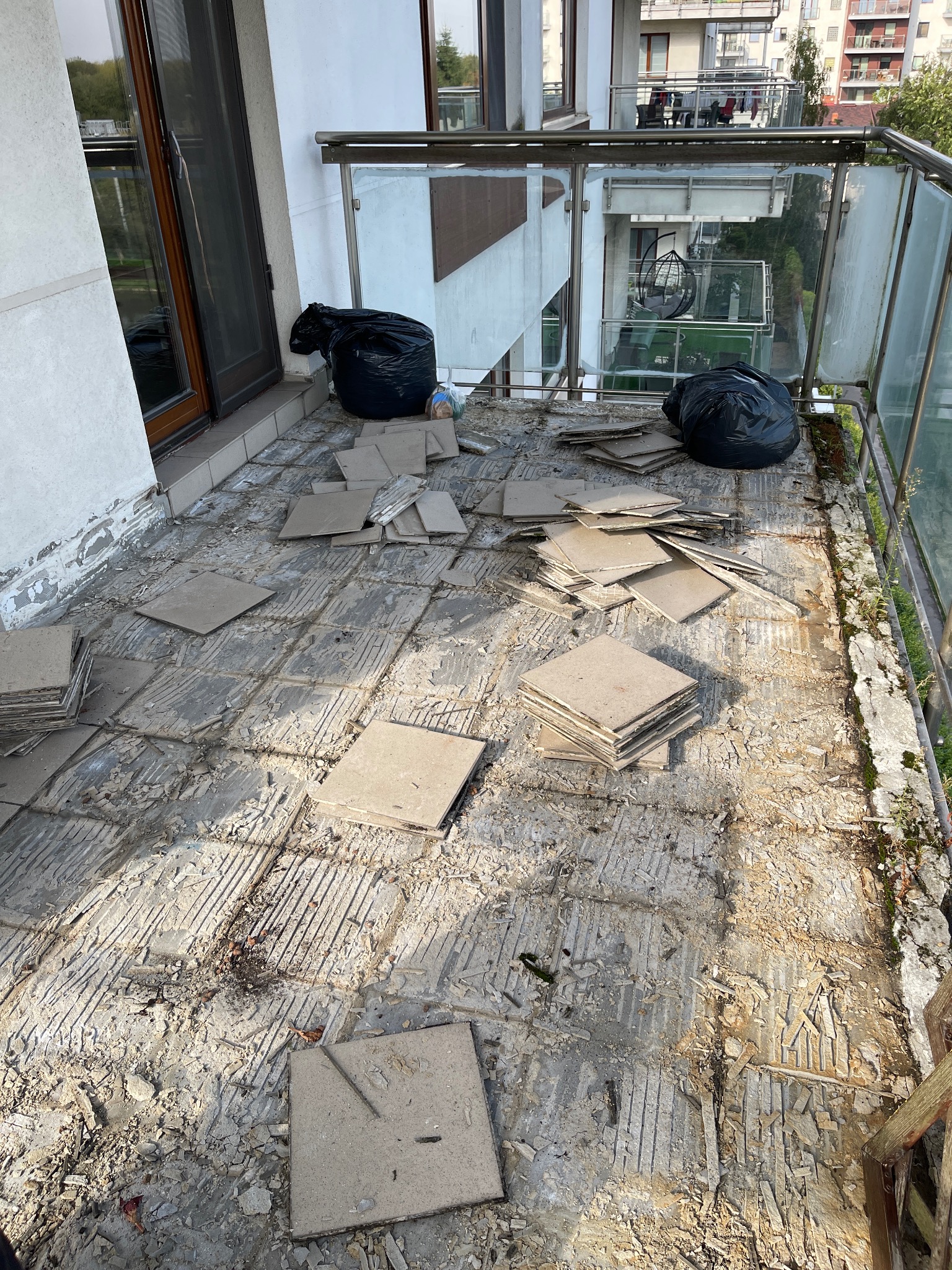 Balkon w trakcie demontażu starych płytek, widoczne pozostałości kleju i gruzu, dwie czarne torby na śmieci z resztkami płytek, balustrada ze szkła i metalu, widok na inne balkony.