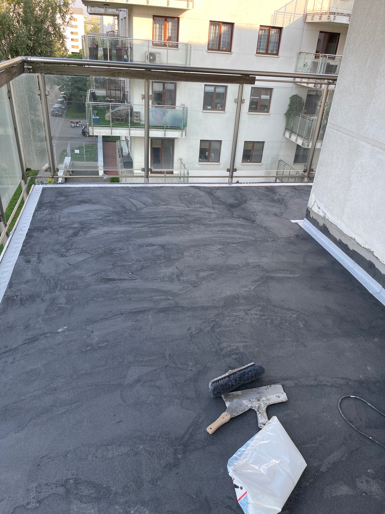 Surowy, szary balkon po wylewce, z widocznymi narzędziami: pędzlem i szpachelką, oraz workiem po zaprawie, na tle budynku mieszkalnego z przeszklonymi balustradami.