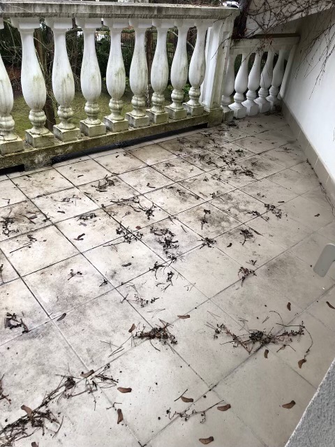 Brudny balkon z balustradą, pokryty liśćmi i gałązkami na kafelkach podłogowych, wymagający sprzątania.