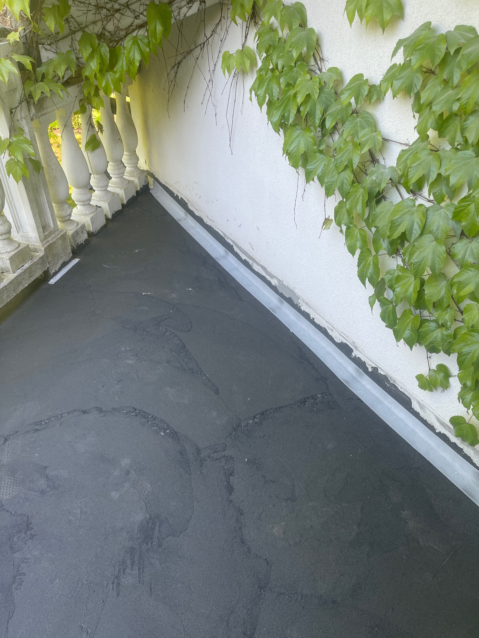 Balkon z nową, czarną hydroizolacją podłogi, widoczne jasne listwy przypodłogowe i pnącza bluszczu na ścianie oraz dekoracyjna balustrada.