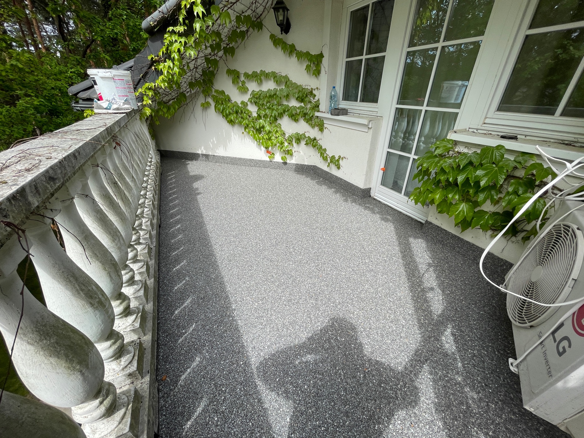 Wykończony balkon z szarą, kamyczkową posadzką, białą balustradą i pnącą się roślinnością na ścianie budynku, widoczny klimatyzator LG