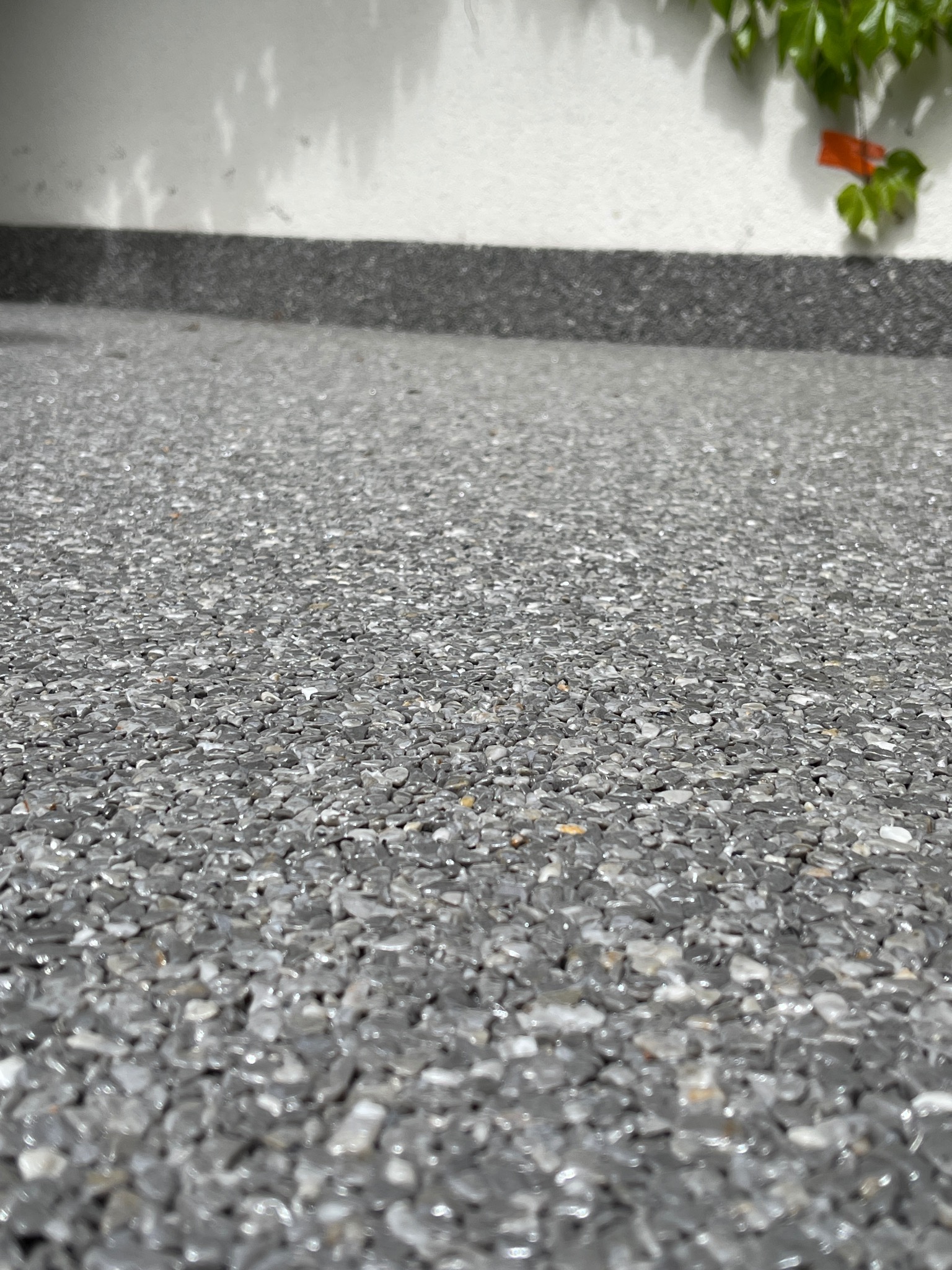 Szara, kamyczkowa posadzka balkonu z widocznym fragmentem białej ściany i rośliny pnącej w tle, wykonana z żywicy epoksydowej.
