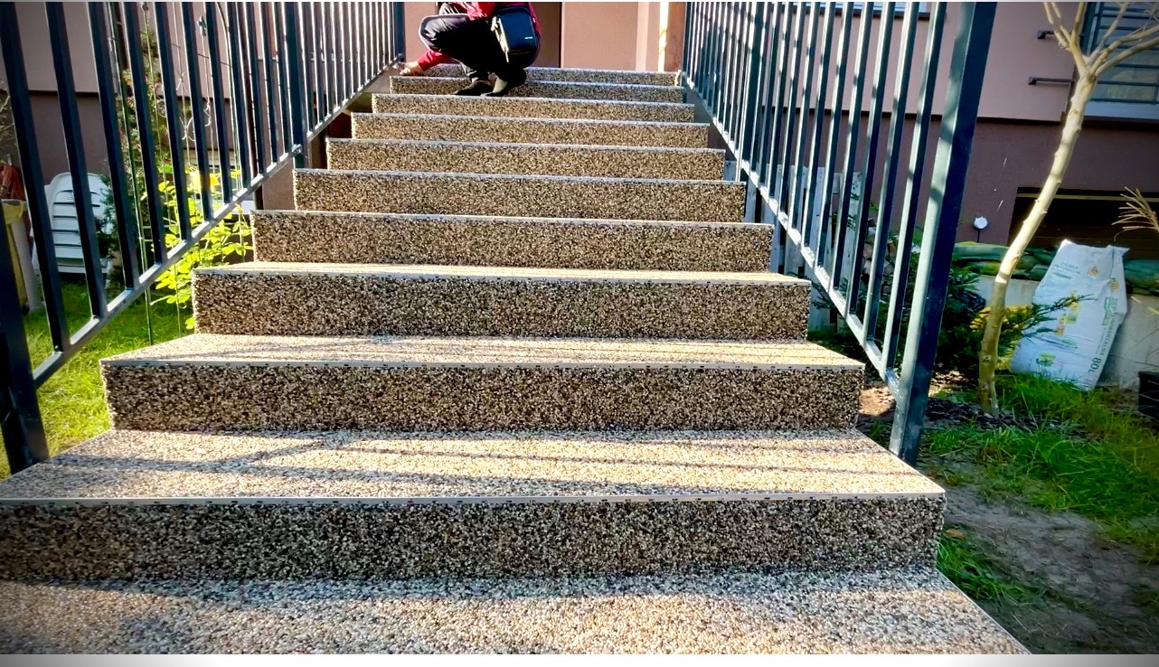 Schody zewnętrzne wykończone drobnym kamieniem w jasnych odcieniach, z czarną metalową barierką, widok z dołu, osoba w górnej części schodów wykonuje prace wykończeniowe.