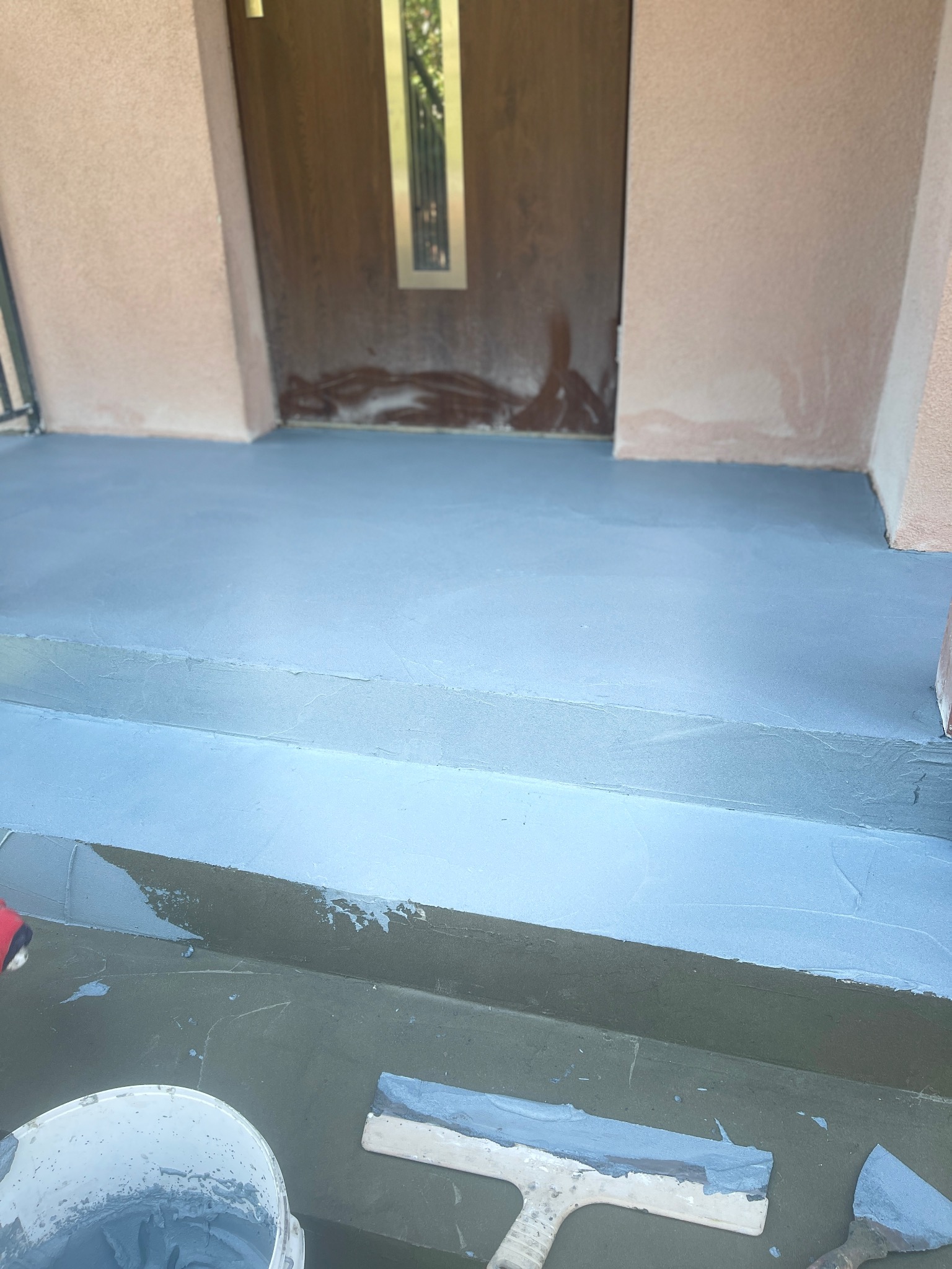 Wyrównywanie szpachlą powierzchni schodów wejściowych do budynku, widoczny kubeł z masą szpachlową i używane narzędzia.