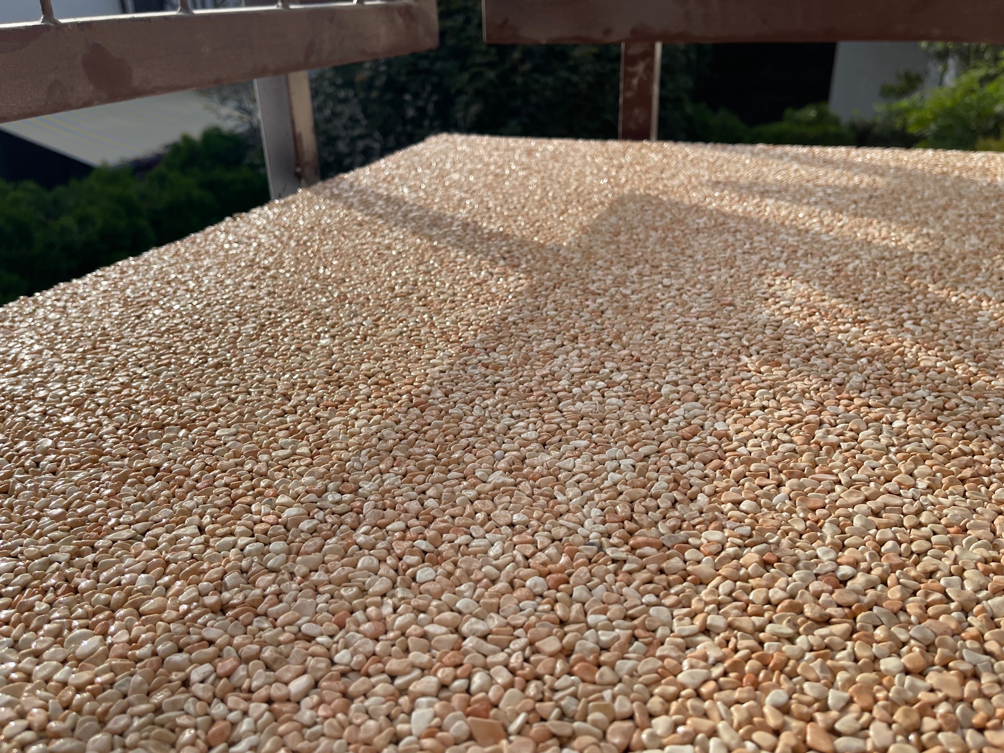 Balkon z wykończeniem z drobnych, jasnych kamyczków, widoczne fragmenty metalowej balustrady i zieleni w tle.