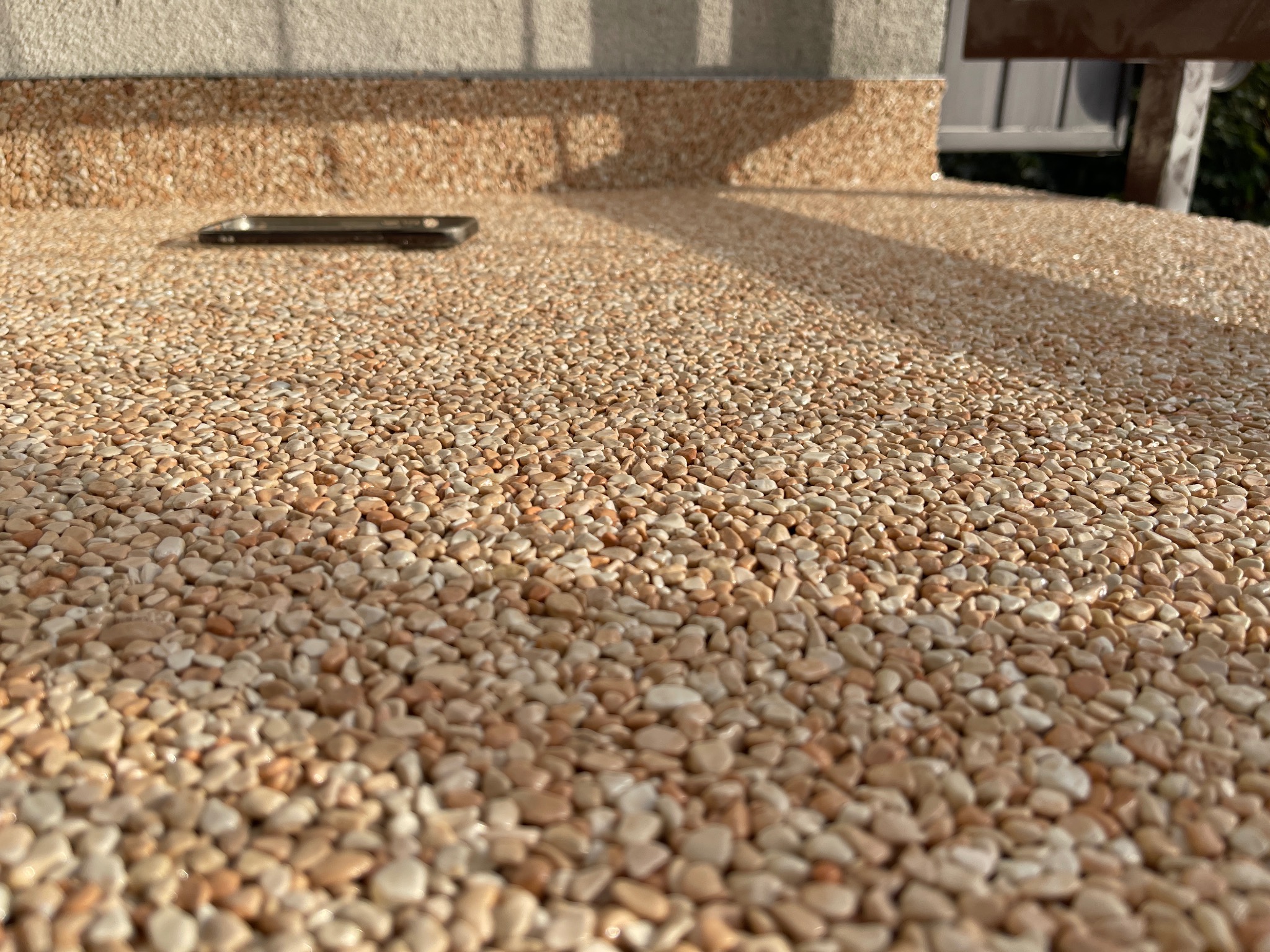 Powierzchnia balkonu wykończona drobnymi, beżowo-brązowymi kamyczkami; na górze leży czarny telefon, a w tle widać fragment ściany i balustrady.