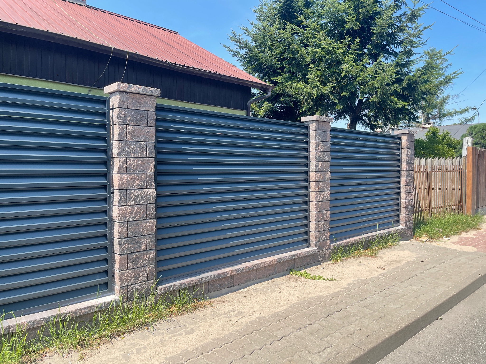 Nowoczesne ogrodzenie z poziomych, grafitowych paneli metalowych, wsparte na słupkach z cegły klinkierowej, widoczne od strony chodnika.