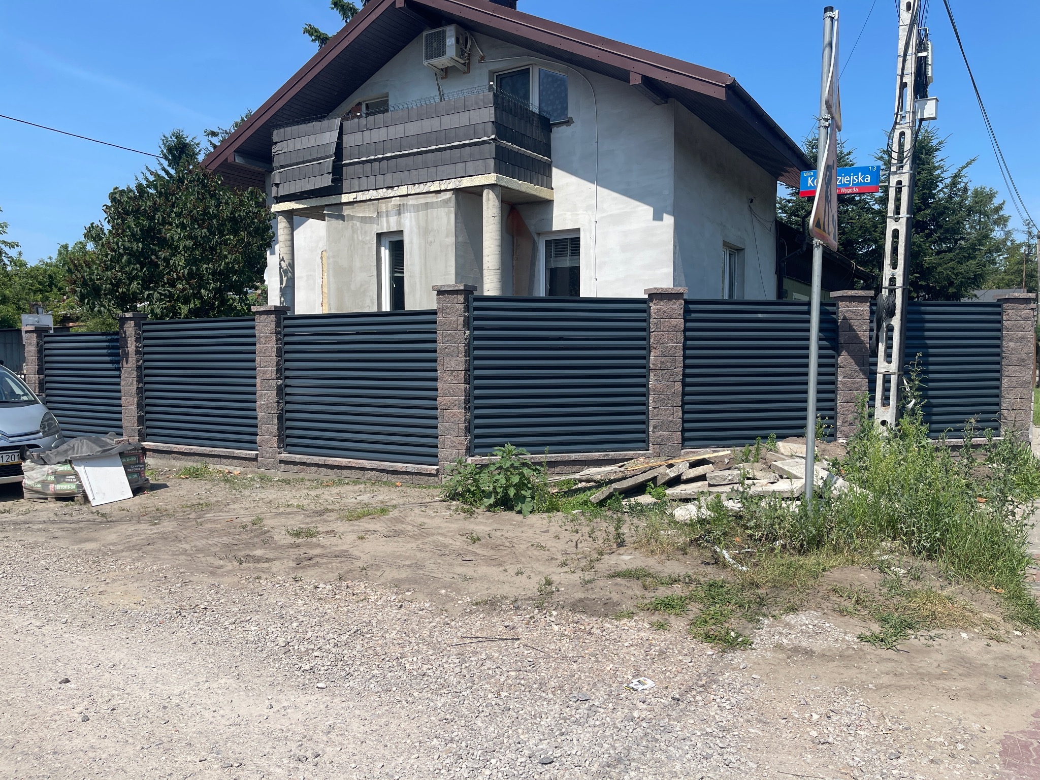 Nowoczesne ogrodzenie z poziomych, grafitowych paneli metalowych i ceglanych słupków, widok z zewnątrz na tle domu z balkonem w trakcie wykończenia, widoczny znak uliczny.