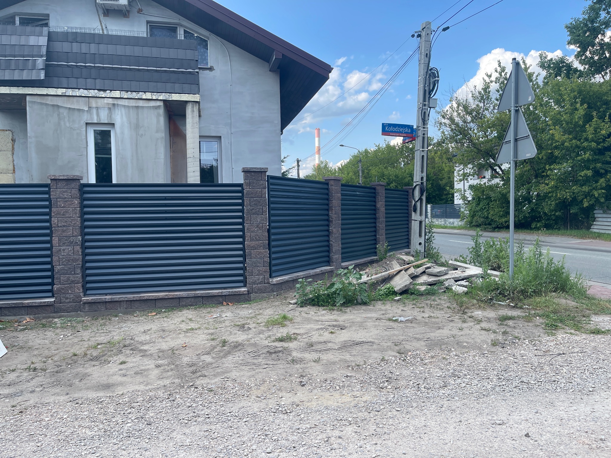 Nowoczesne ogrodzenie panelowe z metalowymi przęsłami i kamiennymi słupkami, widok z zewnątrz posesji, w tle budynek w trakcie wykończenia elewacji.