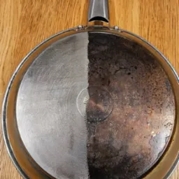 Patelnia z widoczną różnicą przed i po czyszczeniu, połowa powierzchni wyczyszczona do połysku, druga połowa pokryta przypaleniami i zabrudzeniami, leżąca na drewnianym blacie.