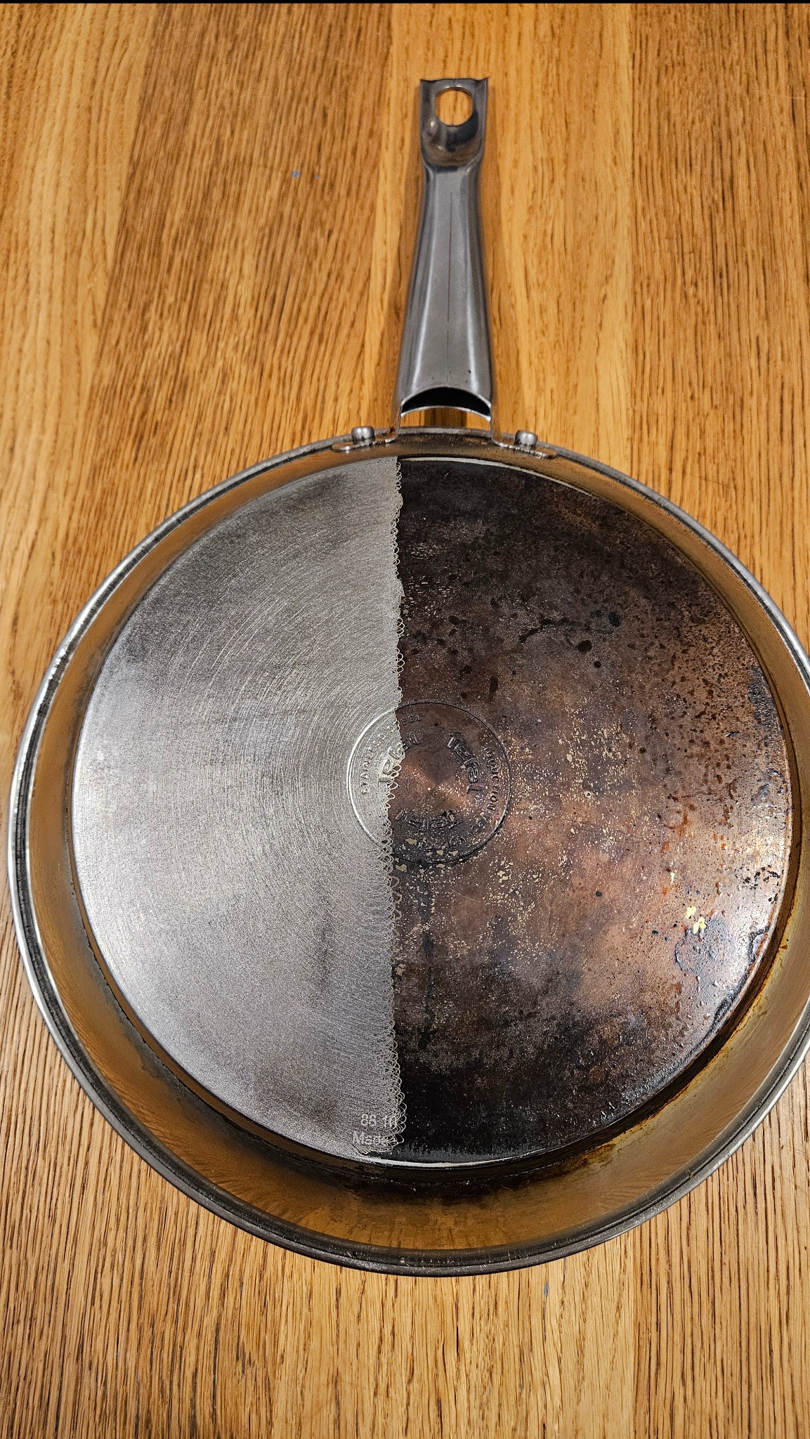 Patelnia z widoczną różnicą przed i po czyszczeniu, połowa powierzchni wyczyszczona do połysku, druga połowa pokryta przypaleniami i zabrudzeniami, leżąca na drewnianym blacie.