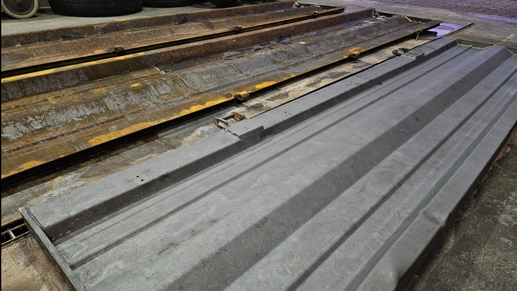 Dwie metalowe formy, jedna oczyszczona z rdzy i pomalowana na szaro, druga w stanie pierwotnym z widoczną rdzą i resztkami farby, leżące obok siebie na betonowej posadzce.