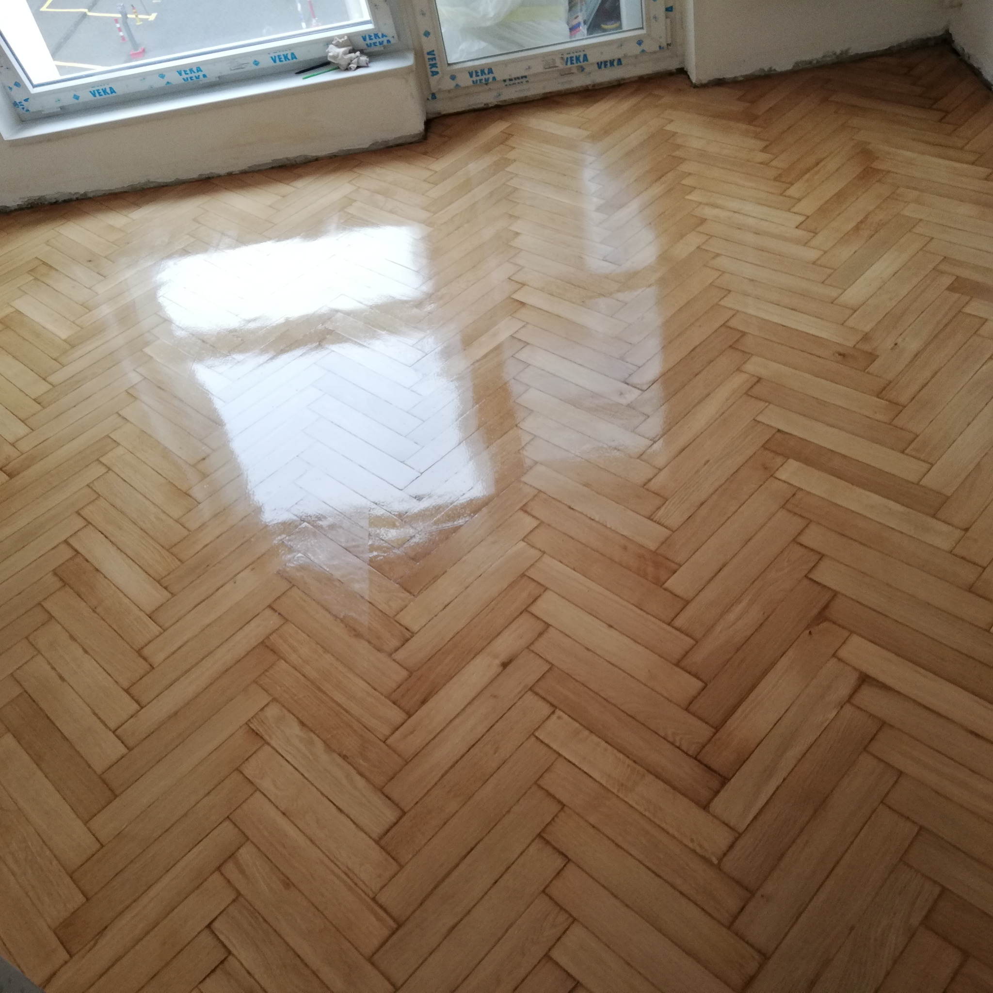 Świeżo polakierowany parkiet ułożony w jodełkę, odbijający światło z okna, widoczne ślady po pracy w postaci drobnych niedoskonałości.
