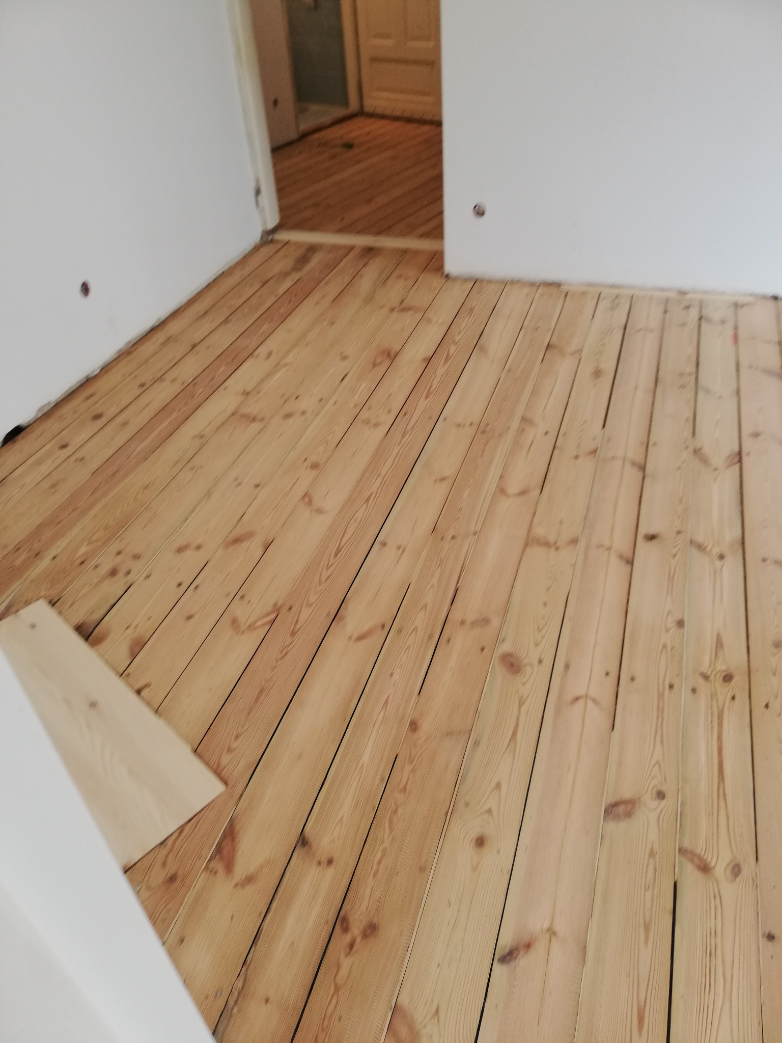 Jasne, drewniane panele podłogowe ułożone w pokoju z białymi ścianami, widoczny fragment nieułożonej deski w rogu.