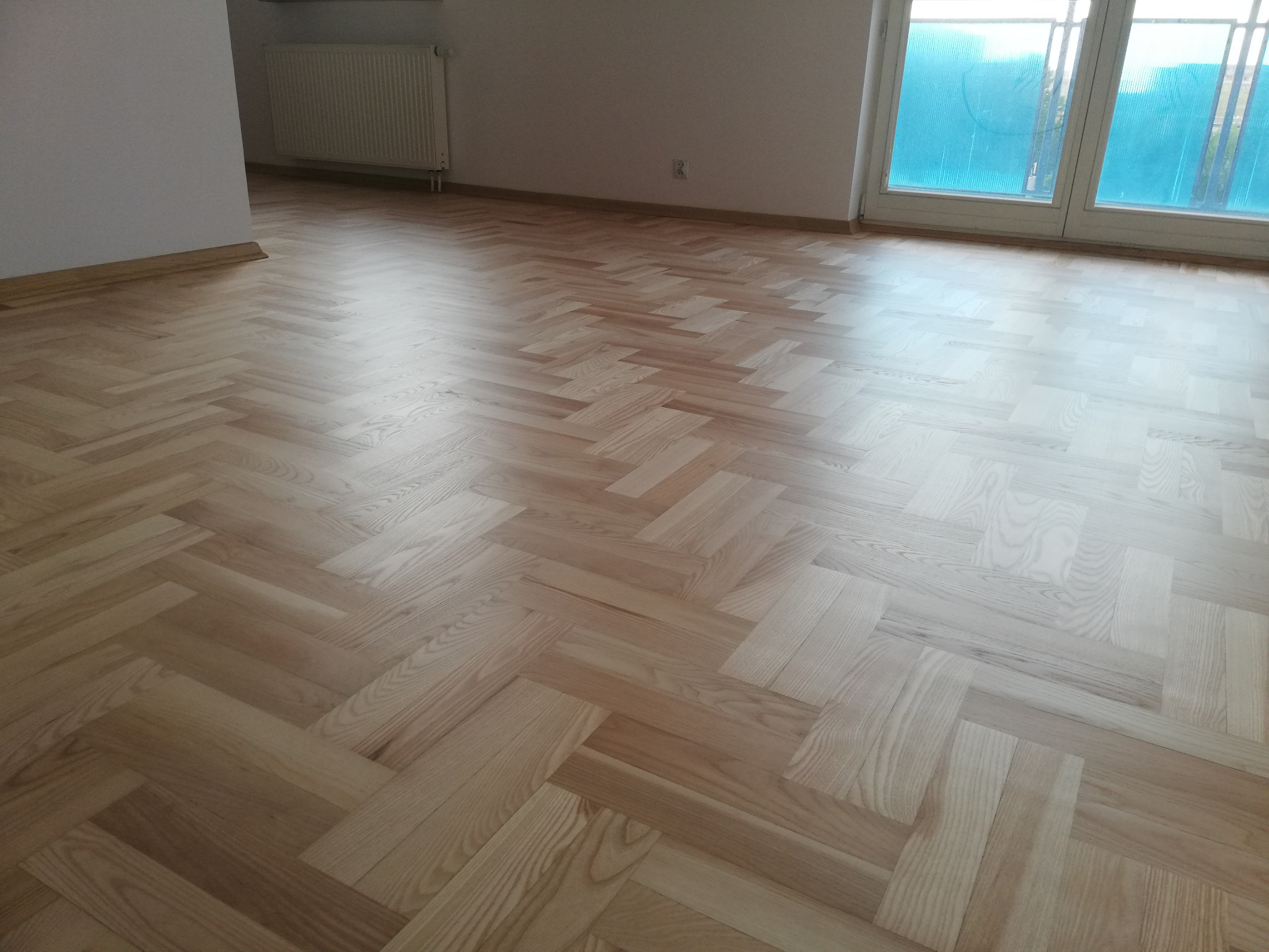 Jasny parkiet ułożony w jodełkę w pustym pokoju z białymi ścianami, grzejnikiem i oknem przesłoniętym niebieską folią.