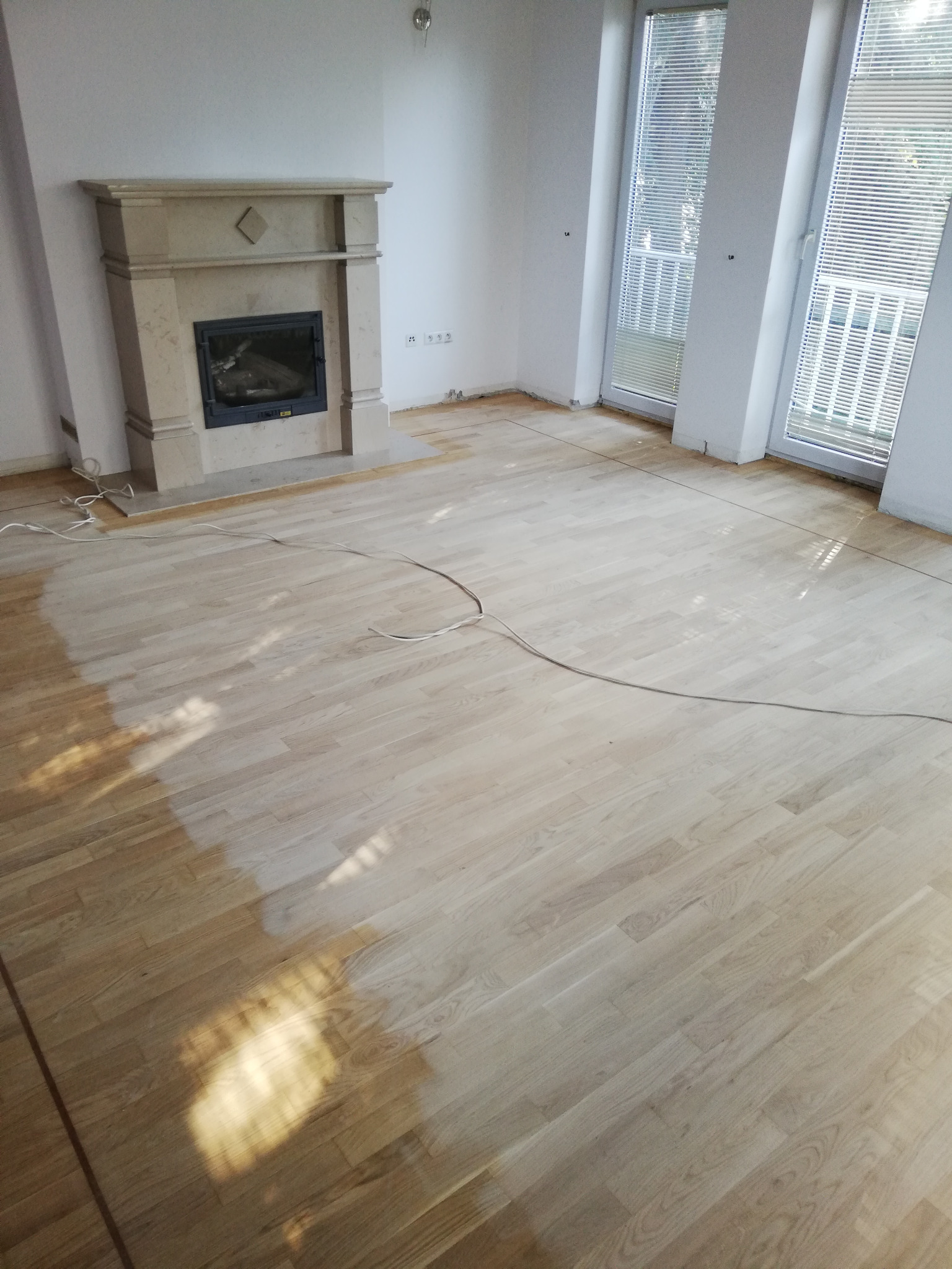 Jasne panele podłogowe ułożone w salonie z kominkiem, widoczne ślady po pracach wykończeniowych przy ścianach i listwach przypodłogowych.