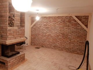 Surowe wnętrze pokoju z ceglanym kominkiem i ścianą z cegły, drewniana konstrukcja wzmacniająca ścianę, parkiet ułożony w jodełkę, widoczne lampy sufitowe