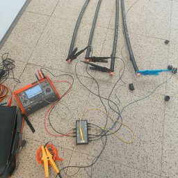 ART MUSIC OBSŁUGA MUZYCZNA IMPREZ LISZEWSKI ARKADIUSZ - Pomiar kabli elektrycznych za pomocą specjalistycznego miernika i akcesoriów na podłodze z płytek. Widoczne kable, miernik, złącza i przewody pomiarowe.