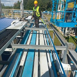 ART MUSIC OBSŁUGA MUZYCZNA IMPREZ LISZEWSKI ARKADIUSZ - Pracownik w kasku i kamizelce na konstrukcji stalowej z kablami, prawdopodobnie podczas prac konserwacyjnych lub montażowych na terenie przemysłowym.