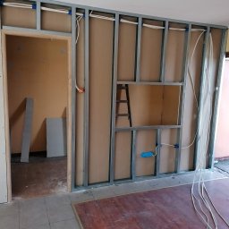 ART MUSIC OBSŁUGA MUZYCZNA IMPREZ LISZEWSKI ARKADIUSZ - Montaż ścianki działowej z instalacją elektryczną wewnątrz. Widoczne kable, metalowa konstrukcja i płyty kartonowo-gipsowe. W tle drabina i fragment pomieszczenia.