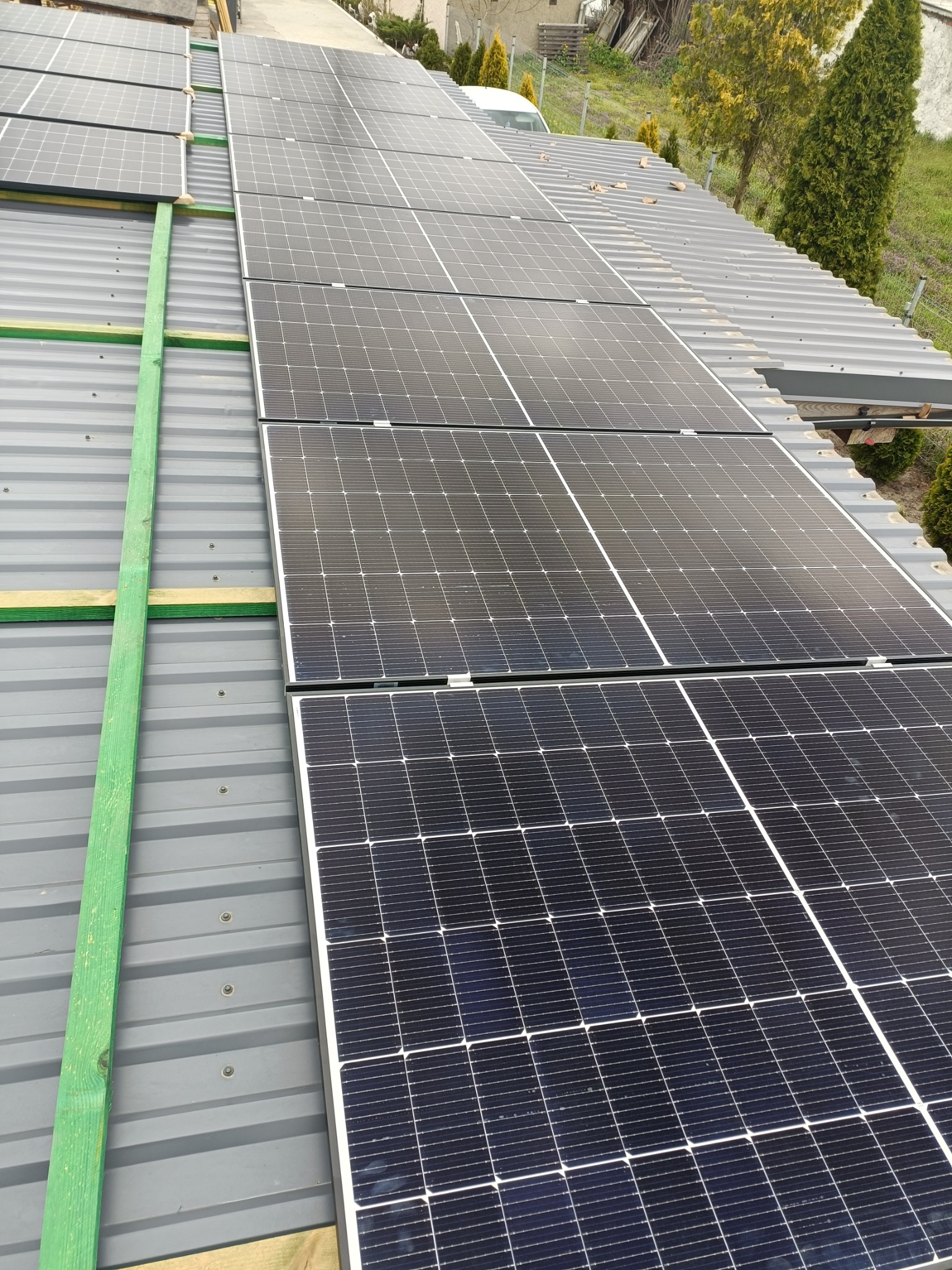 Panele słoneczne na szarym blaszanym dachu, zabezpieczone zielonymi listwami. Widok z góry, na tle drzew i częściowo widocznego budynku.