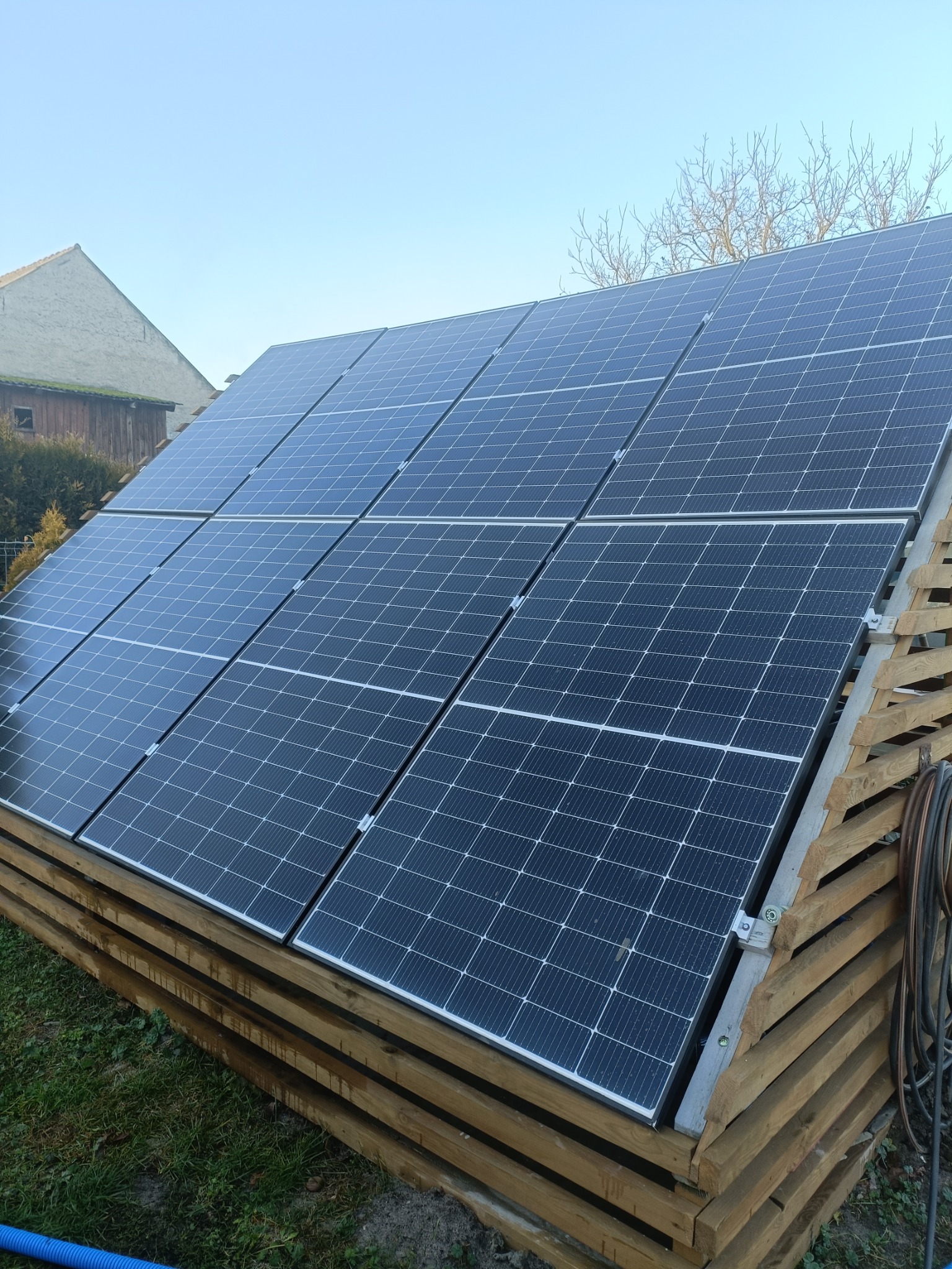 Panel słoneczny na drewnianej konstrukcji w ogrodzie, widok z dołu. Widoczne złącza i kable. W tle budynek i drzewa pod błękitnym niebem.