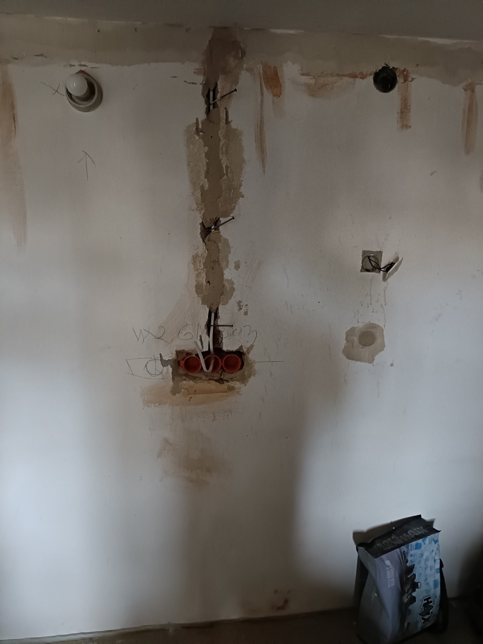 Ściana w trakcie prac elektrycznych: widoczne kable w puszkach, bruzdy w tynku i oznaczenia. Przy podłodze stoi torba z materiałami budowlanymi.