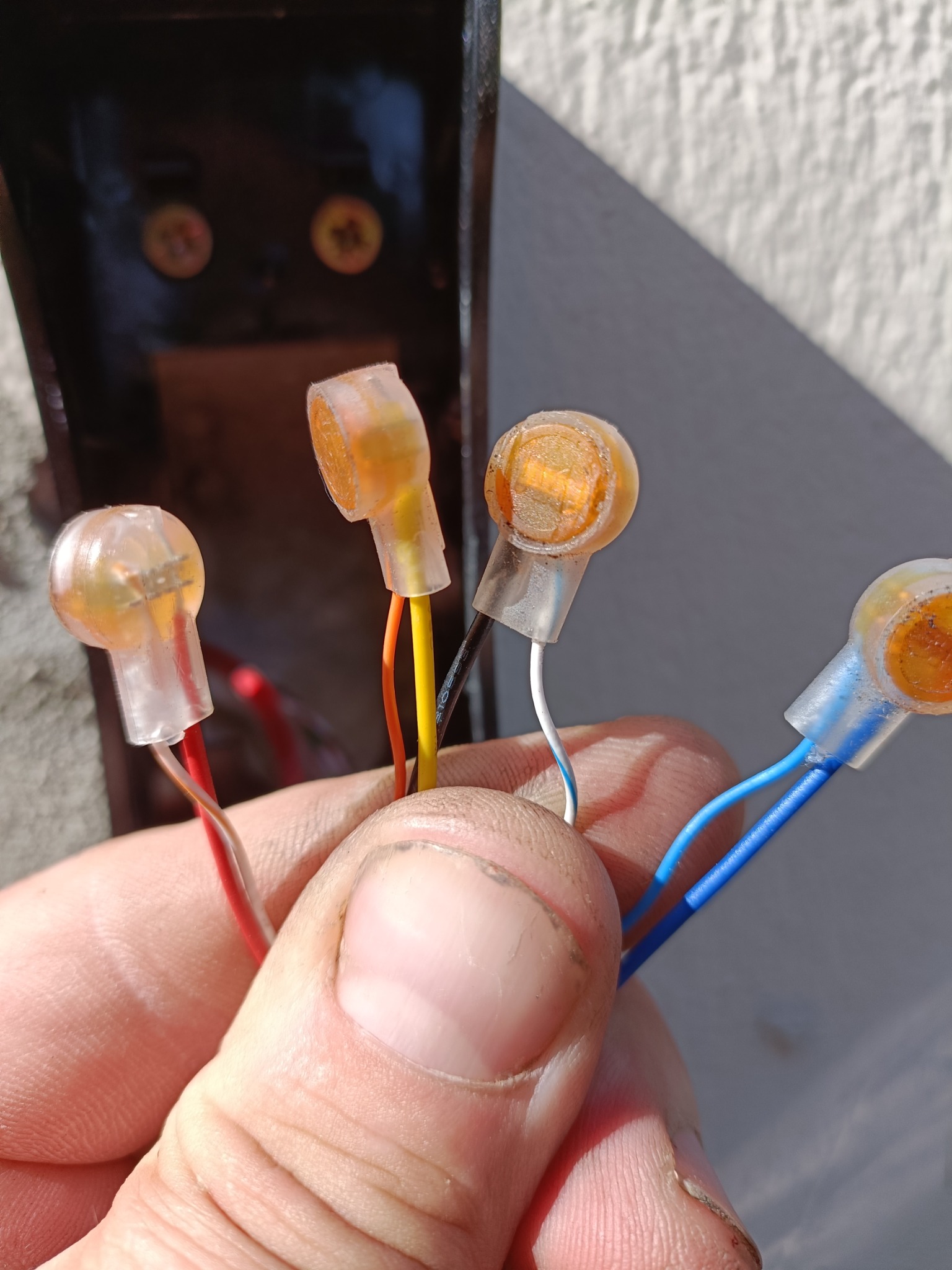 Dłoń trzymająca złączki WAGO z przewodami w różnych kolorach na tle ściany i czarnej obudowy. Widoczne detale połączeń elektrycznych.