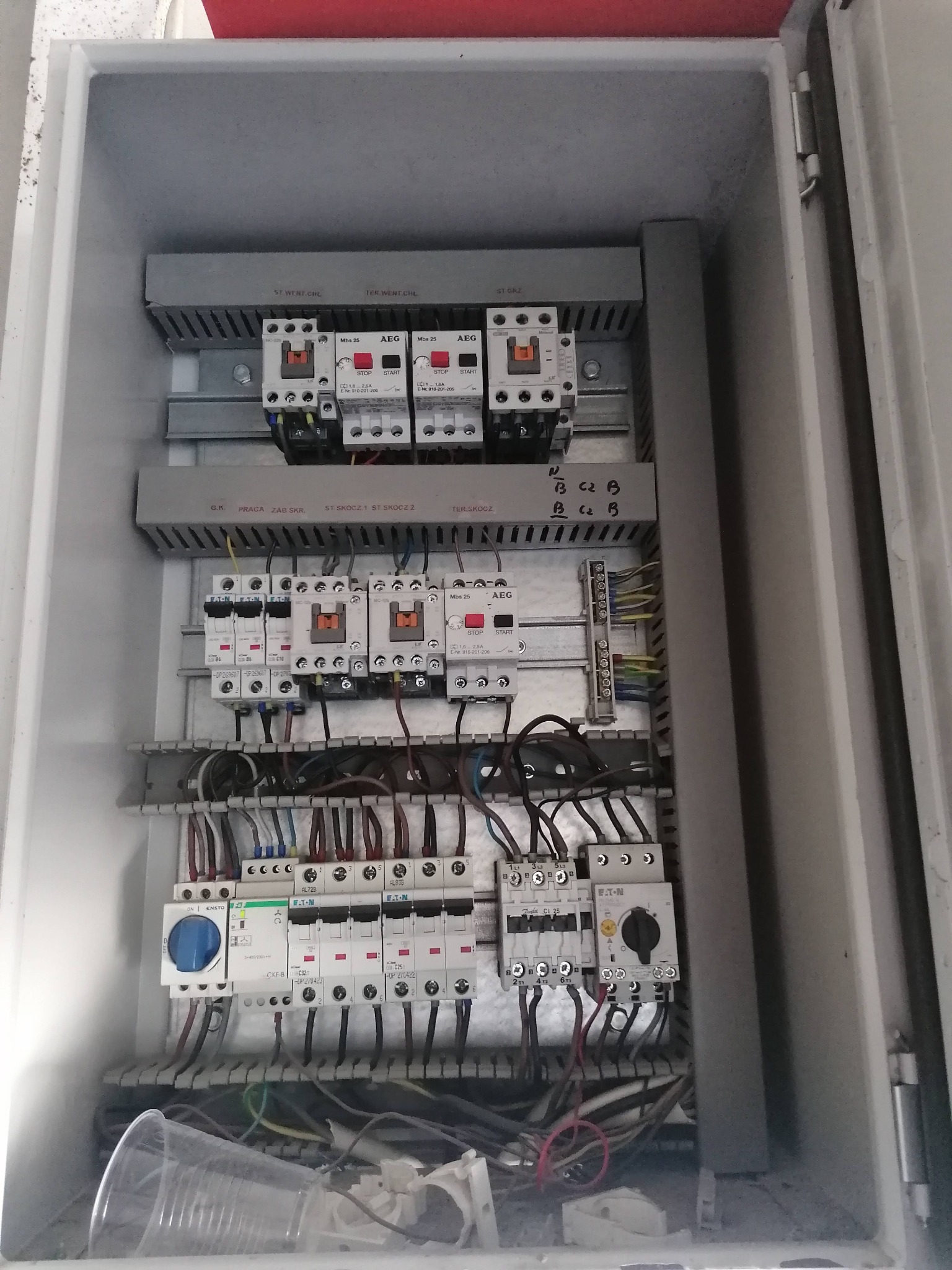 Rozdzielnia elektryczna w obudowie, widoczne zabezpieczenia, styczniki, bezpieczniki i przewody. Elementy AEG i Eaton. W tle plastikowy kubek.