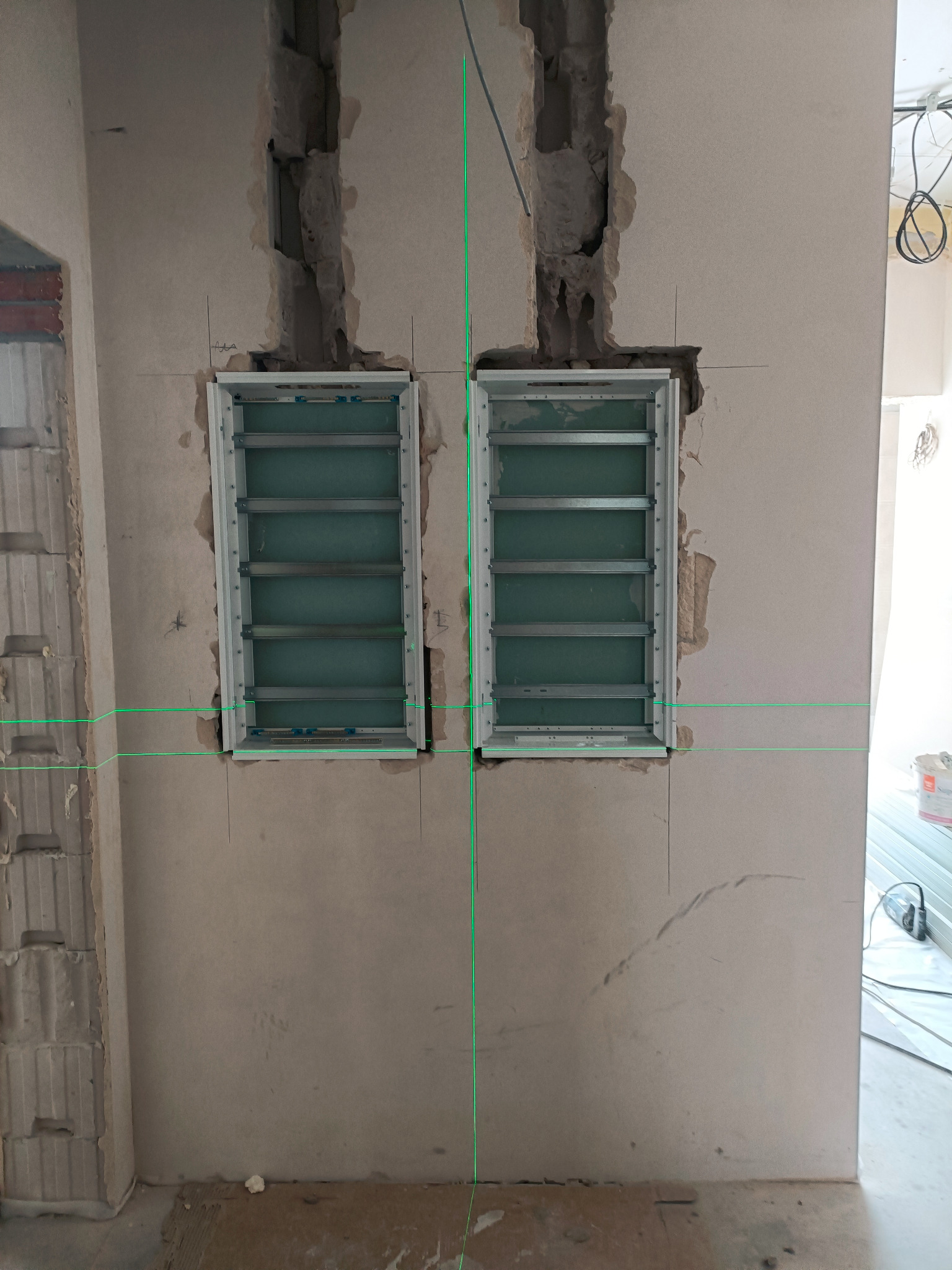 Dwa metalowe panele elektryczne zamontowane w ścianie w trakcie remontu, widoczne przewody i ślady po kuciu. Użycie lasera do poziomowania. Surowy stan wykończenia.