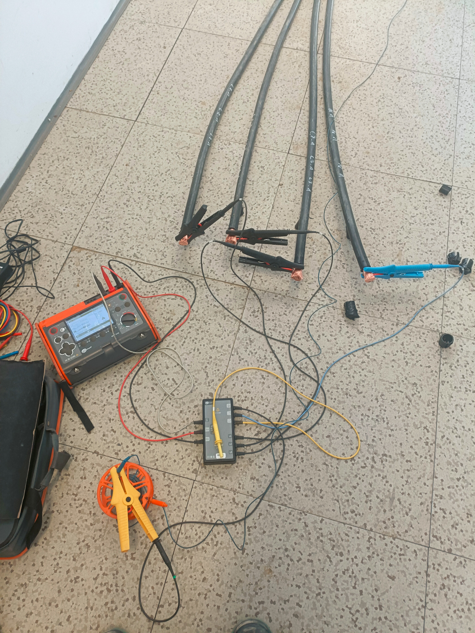 Pomiar kabli elektrycznych za pomocą specjalistycznego miernika i akcesoriów na podłodze z płytek. Widoczne kable, miernik, złącza i przewody pomiarowe.